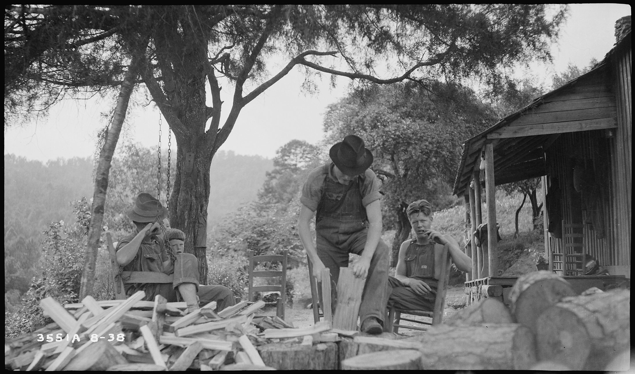 Men of the Dillard Morrow household splitting firewood under a tree beside the log house, children watching