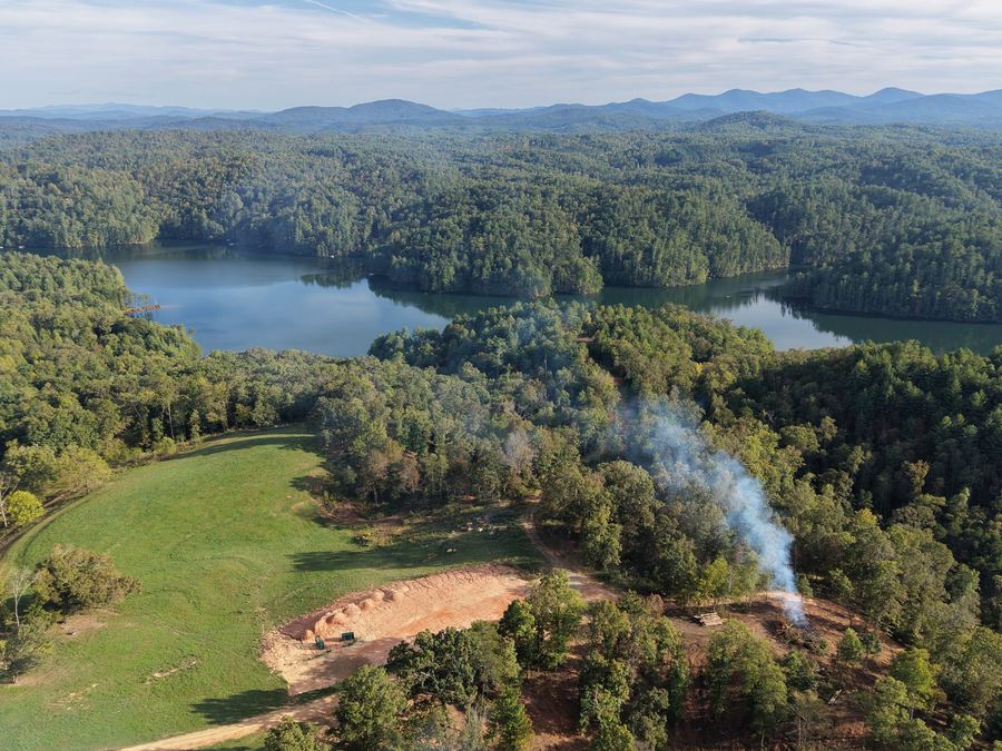 Aerial view of a green pasture, with a thin column of smoke rising from the trees and the lake beyond